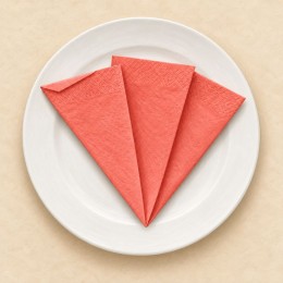 Serviette corail pliée en tulipe à plat : trois volets en éventail et un petit revers, sur une assiette blanche.